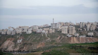 Distant Amman hillside covered with pale apartment buildings above a rocky cut, with a mosque minaret near the skyline.