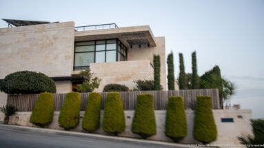 Large modern limestone villa in Amman with broad windows, clipped conical shrubs, cypress trees, and a roadside wall.