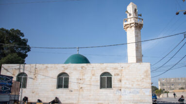 Neighborhood mosque with green dome, white minaret, arched windows, and overhead utility wires.