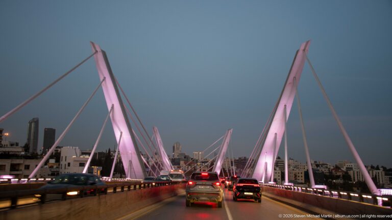 Traffic crossing Amman’s Wadi Abdoun cable-stayed bridge at dusk beneath tall pink-lit pylons and diagonal cables.