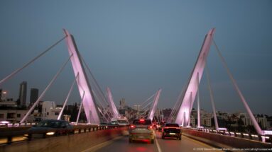 Traffic crossing Amman’s Wadi Abdoun cable-stayed bridge at dusk beneath tall pink-lit pylons and diagonal cables.