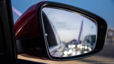 Car side mirror reflecting evening traffic and a bridge in Amman, with Arabic warning text along the lower edge.