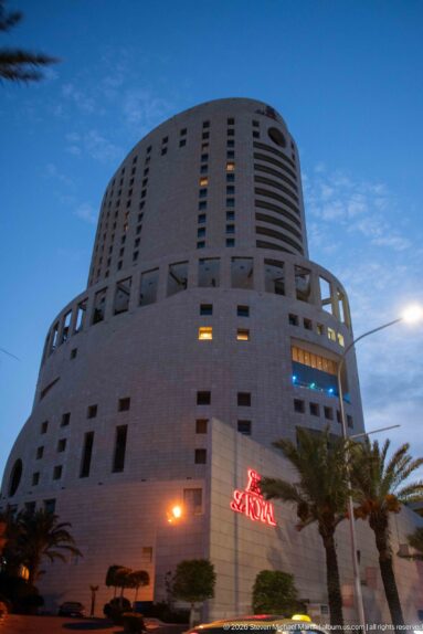 Le Royal Amman at dusk, a rounded stone-clad high-rise with horizontal window bands, red signage, and palm trees.