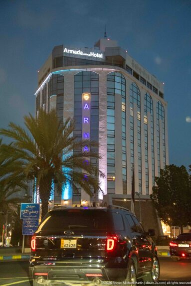 Armada Hotel in Amman at night with illuminated name signs, curved glass façade, palm trees, and cars in front.