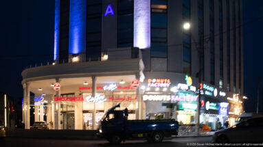 Illuminated Amman corner building with restaurant signs, vertical façade lighting, and a pickup truck passing in front.