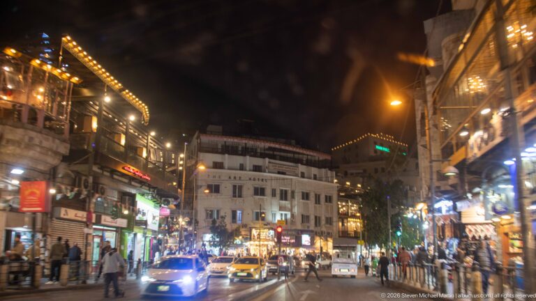 Busy Amman street at night with pedestrians, cars, bright storefronts, signs, and mixed urban lighting.