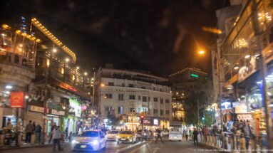Busy Amman street at night with pedestrians, cars, bright storefronts, signs, and mixed urban lighting.