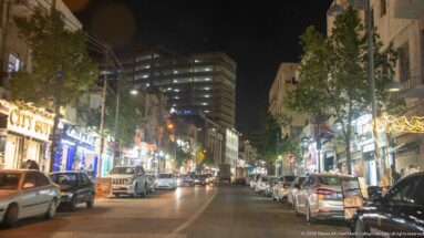 Nighttime Amman commercial street with parked cars, moving traffic, illuminated shops, restaurants, and mid-rise buildings.