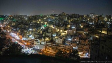 Wide night panorama of Amman with dense hillside housing and a brightly lit road along the lower slope.