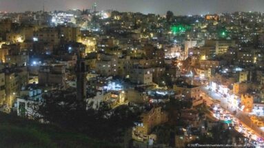 Elevated night view over dense Amman hillside neighborhoods with a brightly lit road running through the valley.