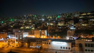 Night cityscape of Amman with dense illuminated housing climbing a steep hillside above larger buildings in the foreground.