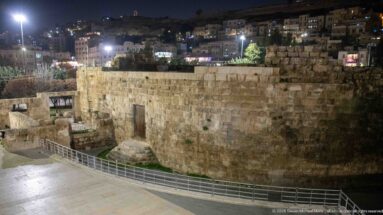 Large ancient stone wall above a lower archaeological area, with a railed walkway and illuminated hillside buildings behind.
