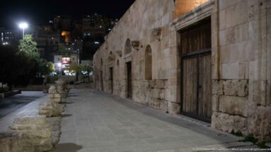 Oblique night view along an ancient stone wall with arched wooden doorways, fragments beside the walkway, and city lights behind.