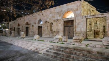 Long ancient stone façade at night with repeated arches, wooden doors, steps, and scattered column fragments.