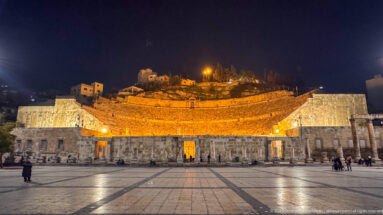 Frontal night view of Amman’s Roman Theater with illuminated seating, long stone stage building, and paved plaza.