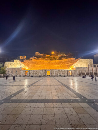 Wide night view across a stone plaza toward the illuminated Roman Theater, with seating rising against the dark hillside.