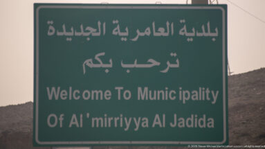 Green bilingual sign reading “Welcome To Municipality Of Al 'mirriyya Al Jadida” with dry hills behind.