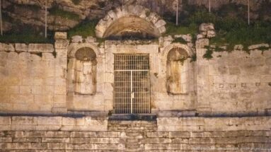 Ancient stone façade at night with a gated central arch, smaller arched niches, carved masonry, and vegetation above.