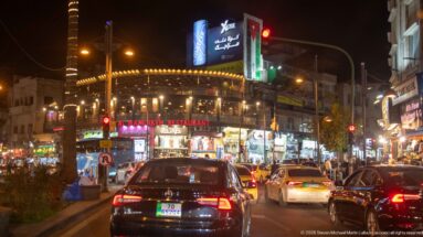 Night traffic at a brightly lit Amman commercial intersection with restaurants, Arabic signage, cars, and pedestrians.