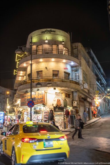 Yellow taxi passes a rounded corner building with shops and pedestrians on an Amman street at night.