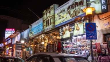 Brightly lit antique and gallery shops line an Amman street at night with hanging lamps and parked cars.