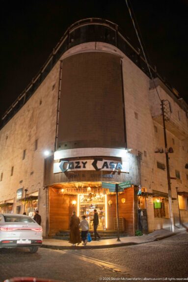 Cozy Casa storefront glowing beneath a rounded stone building as cars and pedestrians pass at night.