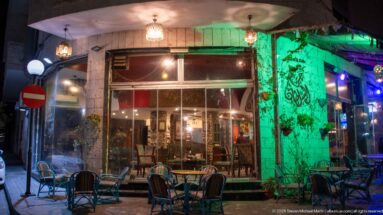 Outdoor café tables and woven chairs beside a green-lit stone wall on an Amman street at night.