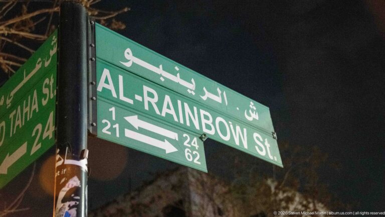 Green bilingual road sign reading Al-Rainbow St in Arabic and English at night.