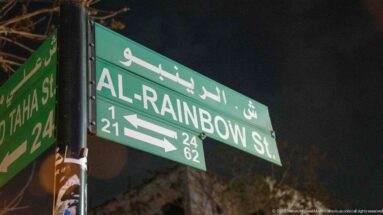 Green bilingual road sign reading Al-Rainbow St in Arabic and English at night.