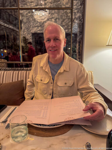 Smiling diner holds a large open menu at a restaurant table with mirrored panels behind.