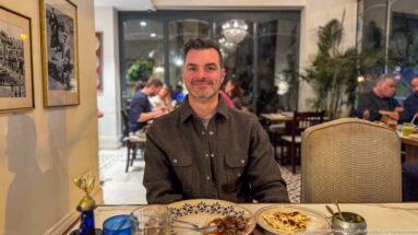 Smiling diner seated at a restaurant table with patterned plates, shared dishes, and other guests behind.