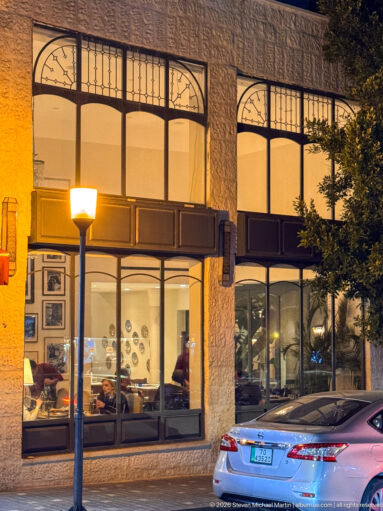 Warm-lit restaurant behind large arched windows in a stone façade, with a streetlamp and parked car outside.