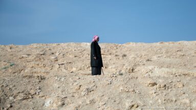 Man in a red-and-white keffiyeh and long dark coat standing alone on pale rocky ground under blue sky.