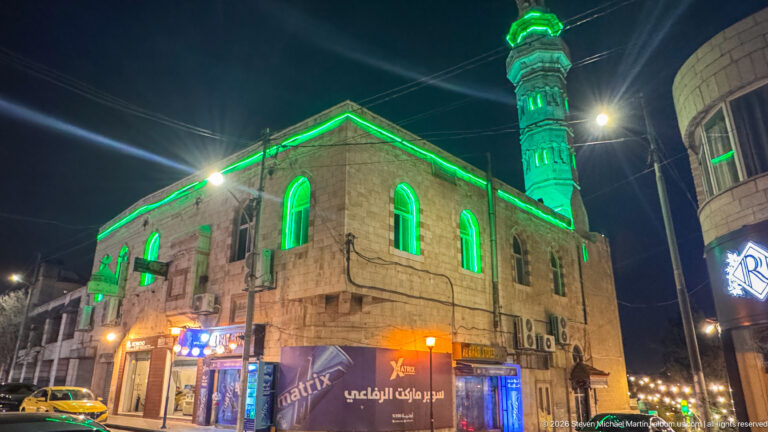 Stone mosque with green-lit arched windows and minaret on an Amman street at night.