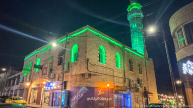 Stone mosque with green-lit arched windows and minaret on an Amman street at night.