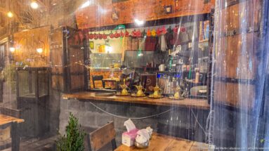 Rain-streaked clear enclosure reveals warm lights, tables, and a small café or restaurant interior at night.