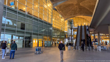 Passengers approach paired escalators in a warm-lit, glass-walled concourse at Queen Alia International Airport.