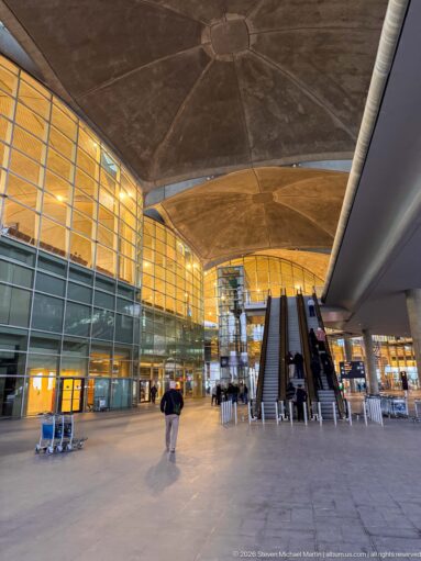 Escalators rise through a glass-walled airport concourse beneath broad concrete roof forms.