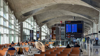 Flight-information screens rise above rows of seated travelers in Queen Alia International Airport’s departure hall.