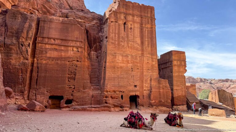 Decorated camels rest beside tall rectangular rock-cut sandstone monuments under a blue sky at Petra.