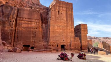 Decorated camels rest beside tall rectangular rock-cut sandstone monuments under a blue sky at Petra.