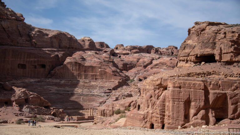 Wide Petra valley view with theater seating at left, tall carved façades at right, and visitors below.