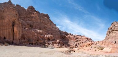 Wide sandy valley at Petra between tall sandstone cliffs, with rock-cut façades and chambers along the left wall.