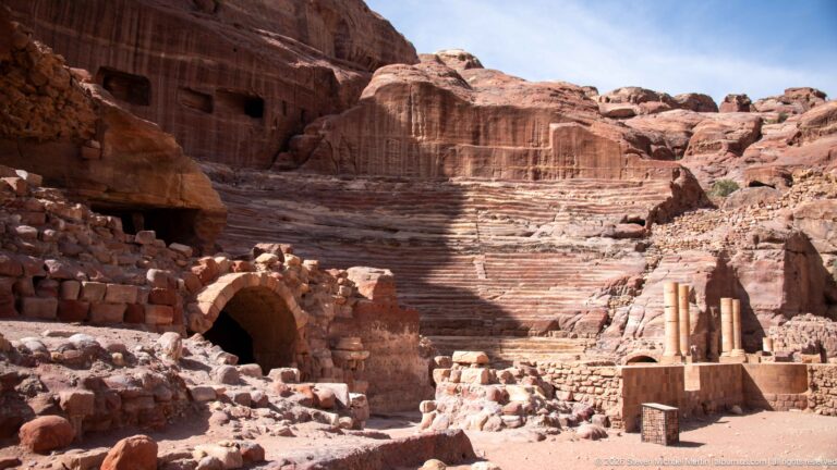 Broad view of Petra’s Theater with rock-cut seating, masonry arch, columns, and surrounding sandstone ruins.