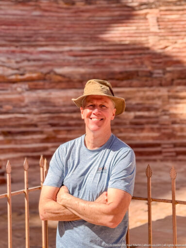 Smiling visitor in a sun hat stands before the blurred sandstone seating tiers of Petra Theater.