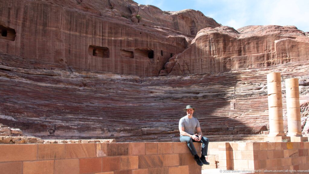Rock-cut stepped seating and stage area of Petra’s Theater, with a visitor seated among the sandstone remains.