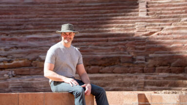 Smiling visitor in a gray shirt and brimmed hat seated on sandstone in front of broad carved rock steps.