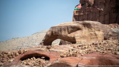 Rounded sandstone outcrop with a natural arch-like opening and cavities, below a cliff where a Jordanian flag is visible.