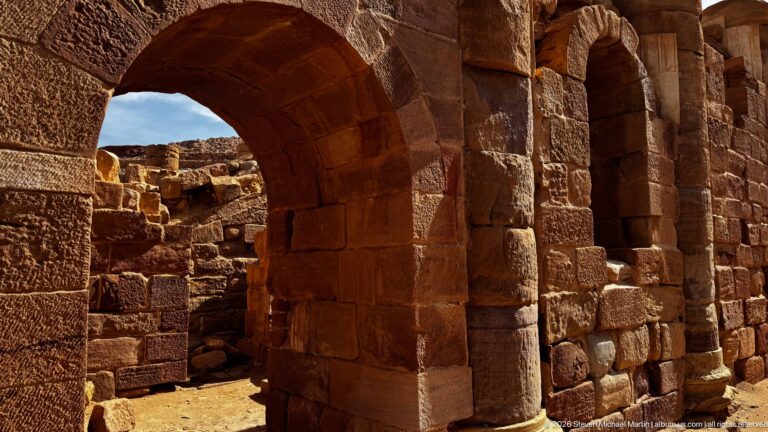 Two heavy semicircular sandstone arches and thick block walls forming a shadowed passage through archaeological ruins.