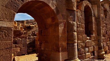 Two heavy semicircular sandstone arches and thick block walls forming a shadowed passage through archaeological ruins.
